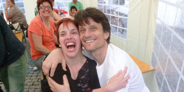 Ein Mann umarmt eine Frau. Beide lachen zum Fotografen, laut mit offenem Mund und strahlenden Augen. Im Hintergrund fröhliche Menschen an Festtischen.