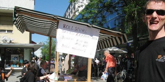 Ein Plakat an einem Marktstand kündigt Rollstuhlselbsterfahrung an. Mehrere RollstuhlfahrerInnen sind am Stand beschäftigt. Leere Rollstühle stehen bereit. Ganz rechts ein Mann mit Sonnenbrille lädt ein zum Sitzen.