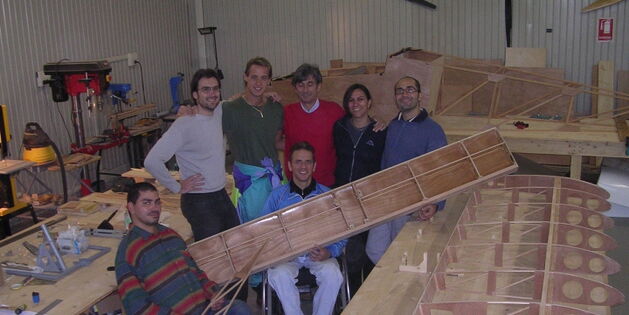 Eine Werkstatt. Das Holzgerippe eines Flugzeugflügels liegt auf dem großen Arbeitstisch im Vordergrund. Dahinter präsentieren sich Mario Pozzini mit seinem Team. Zwei Rollstuhlfahrer zeigen hölzerne Bauteile des Flugzeugs.