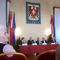 Die Referenten am Podium im Wappensaal des Wiener Rathauses. Der Österreichische Adler über den Köpfen, Fahnen rechts und links.