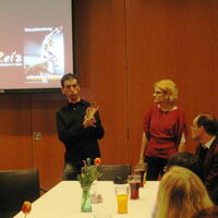 Angela Kreil und Andreas Guth moderieren vor einem weißen mit Getränken und Blumen gedeckten Tisch. Im Hintergrund die Leinwand mit dem Begrüßungsbild der Reiz-Sexualberatung.
