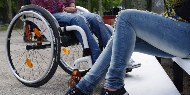 Zwei Beinpaare im Gespräch? Ein Beinpaaar in Jeans mit blauweißen Turnschuhen. Angewinkelt in Sitzposition auf weißen Stufen. Ihm zugewandt das andere Beinpaar. Blaue Jeans, hellgraue Turnschuhe, im Rollstuhl mit weißem Rahmen und schwarzen Reifen.