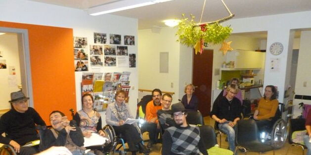 Mensch Zuerst Vorarlberg und Reiz bei der Jahressitzung im Dornbirner Büro. Sechs gutgelaunte RollstuhlfahrerInnen sind auf der linken Bildhälfte versammelt. Rechts davon die StuhlbesitzereInnen. Zwei Hände mit Fotokamera schauen ins Bild hinein.