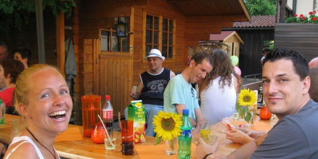 Lachende Frauen und Männer sitzen an Biertischen. Durstlöscher stehen bereit zur Kühlung. Sonnenblumendekoration. Unter einem Tisch lacht ein Hund in die Kamera.