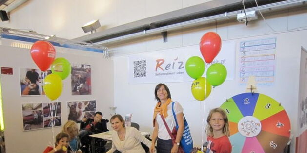 Der Messestand von Reiz mit dem bunten Glücksrad. Kinder mit Luftballons lachen in die Kamera. In der Mitte eine Frau mit selbstbewusstem Blick. Eine Rollstuhlfahrerin lehnt sich zu zwei Schülern, die ins Bild streben.