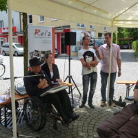 Obmann Andreas Guth bei der Eröffnungsrede. Reinhard Zischg übersetzt. Marianne Schulze lacht fröhlich. Auf dem Kopfsteinpflaster reflektiert die Sonne.