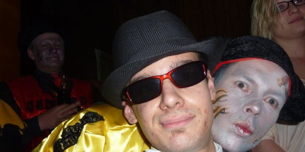 Mann mit Sonnenbrille und Hut lächelt cool in die Kamera. Hinter ihm schaut eine Maske mit offenen Augen und Kussmund hervor.
