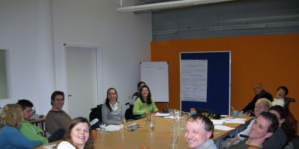Das Bild zeigt den Büroraum in Bludenz mit der Reiz-orangen Wand im Hintergrund. Die Teams von Mensch Zuerst-People First Vorarlberg und Reiz sitzen um einen großen Tisch. Alle schauen fröhlich in die Kamera.