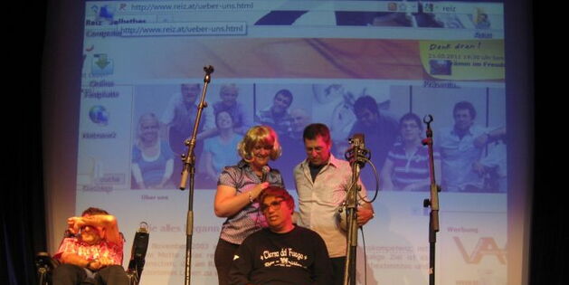 Der Spenden-Scheck wurde übergeben. Er lehnt an den Unterschenkel eines Rollstuhlfahrers. Ein anderer Rollstuhlfahrer hält sich entsetzt die Hand vor die Augen. Eine Frau und der Spender stehen befriedigt hinter dem "Scheckhalter".