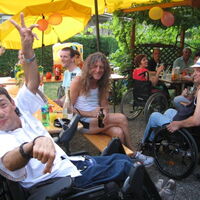 Gelbleuchtende Sonnenschirme. Luftballone. Biertische und Bänke. RollstuhlfahrerInnen in Sommerkleidung lachen zum Betrachter. Zum Wohl beim Sommerfest.