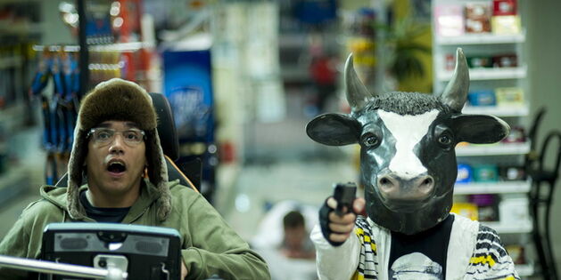 Szene aus dem Film Vielen Dank für Nichts. Ein Mann verkleidet mit einem weiß-schwarzen Faschingskostüm und mit einer Kuhmaske auf dem Kopf hält eine Waffe in der Hand und zielt auf den Betrachter. Der Mann neben ihm trägt eine Pelzmütze auf dem Kopf, sit