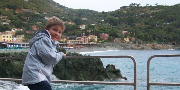 Ein Junge mit kritischem Blick hält sich am Geländer fest. Unter ihm Wellengischt. Im Hintergrund ein Felsen im Wasser, Sandstrand und ein Dorf am ligurischen Meer.