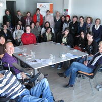 Die 3. Klasse der Krankenpflegeschule Bregenz versammelt zum Schulunterricht im Reiz-Büro. Alle sitzen um einen großen Tisch und lachen in die Kamera. Im Vordergrund die Reiz-MitarbeiterInnen Claudia Fessler, Sabrina Nitz, Manuela Nitz und Süleyman Kurt.