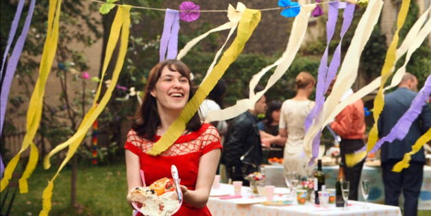 Eine junge Frau in rotem Kleid trägt mit glüchlichem Lachen ihren Teller voll Kuchen zwischen bunten Girlanden. Im Hintergrund gedeckte Tische und Partygäste einer Gartenfeier. Fotoquelle: Alamode