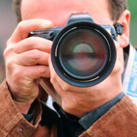 Foto: zoonar. Ein Mann hält eine Fotokamera vor seinem Gesicht direkt zum Betrachter gerichtet, bereit ein Bild zu knipsen.