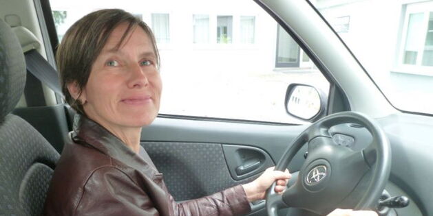 Eine junge Frau sitzt in einem Fahrzeug am Steuer und hält das Lenkrad sicher mit beiden Händen. Ihre blauen Augen lächeln in die Kamera.