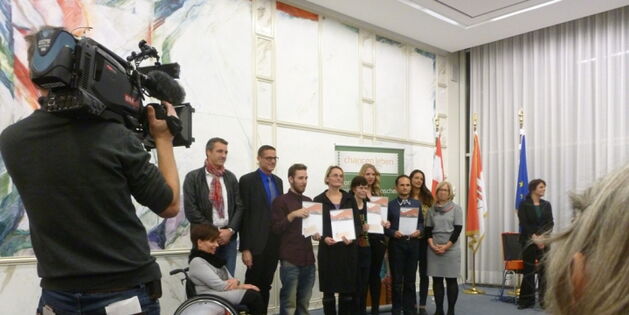 Links im Bild ein ORF-Kameramann mit dem Rücken zum Betrachter, Kamera geschultert und auf die PreisträgerInnen gerichtet. Die fünf PreisträgerInnen stehen in der Mitte und zeigen ihre Urkunden. Landesrat Bernhard und Jurymitglieder umrahmen die Sieger.