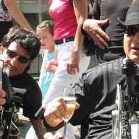 Menschenkörpergewimmel. Im Vordergrund Reinhard sonnenbebrillt und schwarz gekleidet mit gefülltem Sektglas in der Hand. Eine Hand mit gefülltem Sektglas nähert sich vom linken Bildrand zum Anstoßen. Sülo beobachtet die Szene.