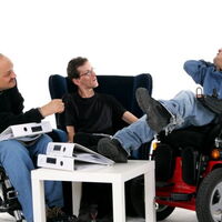 Drei Männer im Gespräch. Zwei Männer wenden sich einem Rollstuhlfahrer zu, der auch mit Händen und Füßen redet. Zwischen ihnen ein Tisch mit Antenordnern.