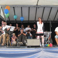 Die Band C.C.Mate mit VertreterInnen des Vorarlberger Krüppelkabaretts auf der Bühne. Bunte Ballone schmücken den linken Rand der Bühne.