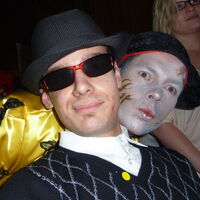 Mann mit Sonnenbrille und Hut lächelt cool in die Kamera. Hinter ihm schaut eine Maske mit offenen Augen und Kussmund hervor.