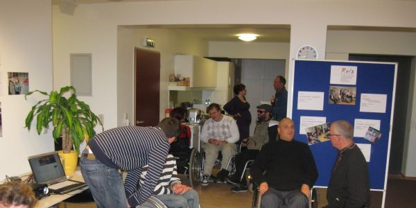 Mehrere Menschen im Rollstuhl, vertieft in Gespräche. Im Zentrum beugt sich ein Assistent zu einem Rollstuhlfahrer. Rechts davon zwei Menschen im Gespräch: einer kniet, Hände in den Hosensäcken, redet; einer sitzt im Rollstuhl, hört aufmerksam zu.