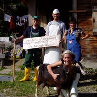 Alppersonal. Im Vordergrund kniet Sennerin Simone König bei der Ziege Brunhilde und umarmt das Tier. Dahinter stehen der Beisenn in weißer Schürze, ein Knecht mit Mistgabel und eine Magd mit Hütestecken. Alle lachen zum Betrachter.