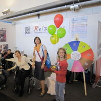Der Messestand von Reiz mit dem bunten Glücksrad. Kinder mit Luftballons lachen in die Kamera. In der Mitte eine Frau mit selbstbewusstem Blick. Eine Rollstuhlfahrerin lehnt sich zu zwei Schülern, die ins Bild streben.