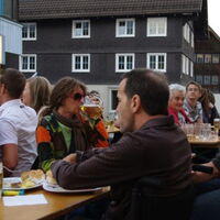 Viele Menschen sitzen entspannt oder fröhlich an Biertischen. Ein Mann und eine Frau im Vordergrund essen Grillspezialitäten. Eine Frau im Hintergrund trinkt aus dem Bierkrug.