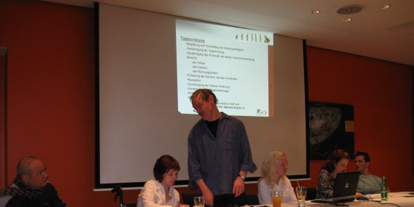 Am Podium links sitzen Reinhard und Julia, die das Protokoll führt. Eberhard steht als Moderator in der Mitte und spricht zum Publikum. Links von ihm sitzen Angi und Andi und lauschen Eberhards Vortrag.
