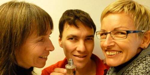 Ein rotbehemdeter Mann hält die Parfumflasche des Duftes Reiz Unisex in der Hand. Zwei Frauen schnuppern daran.