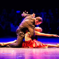 Ein tanzendes Paar umarmt beim Tango. Die Frau in rotem Kleid im Spagatschritt am Boden, Hand elegant gestreckt, ihr rechtes Bein hält das Bein des Tänzers am Boden fest. Der Tänzer kauert auf einem Bein, hält die Frau mit den Armen im Tanz..
