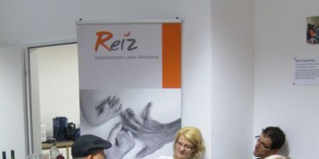 Drei Frauen, zwei Männer, ein Baby im Gespräch. Eine Frau und ein Mann sitzen im Rollstuhl. Ein Hund und eine Blume machen den Vordergrund. Im Hintergrund der Reiz-Banner mit den drei Händen.
