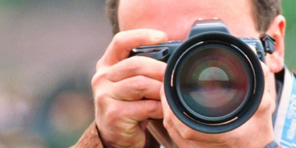 Foto: zoonar. Ein Mann hält eine Fotokamera vor seinem Gesicht direkt zum Betrachter gerichtet, bereit ein Bild zu knipsen.