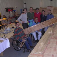 Eine Werkstatt. Das Holzgerippe eines Flugzeugflügels liegt auf dem großen Arbeitstisch im Vordergrund. Dahinter präsentieren sich Mario Pozzini mit seinem Team. Zwei Rollstuhlfahrer zeigen hölzerne Bauteile des Flugzeugs.