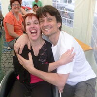 Ein Mann umarmt eine Frau. Beide lachen zum Fotografen, laut mit offenem Mund und strahlenden Augen. Im Hintergrund fröhliche Menschen an Festtischen.