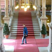 Ein hoher Raum mit gewölbter Decke und Säulenreihe entlang des Stiegenaufgangs. Ein roter Teppich belegt die lange Stiege. Vor der Stiege Andreas Guth dreht sich dem Fotografen zu. Ein Lüster mit vielen Lichtern erhellt den Raum.
