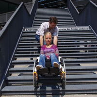 Ein Mann zieht eine junge Frau im Rollstuhl über eine lange Treppe. Die Frau schaut streng in die Kamera.