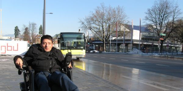 Sueleyman Kurt wartet in seinem Elektrorollstuhl auf den einfahrenden gelben  Landbus. Im Hintergrund blauer Himmel und das Kulturhaus Dornbirn.