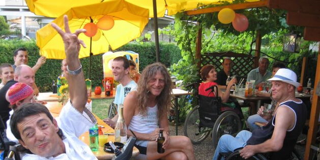 Gelbleuchtende Sonnenschirme. Luftballone. Biertische und Bänke. RollstuhlfahrerInnen in Sommerkleidung lachen zum Betrachter. Zum Wohl beim Sommerfest.