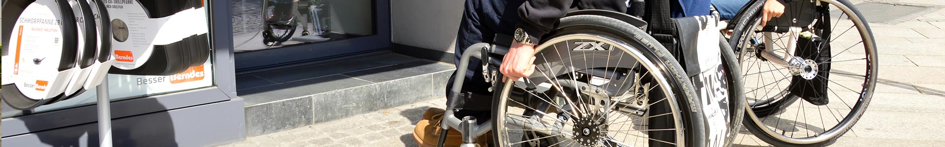 Panoramabild: Zwei RollstuhlfahrerInnen stehen vor einer Stufe.