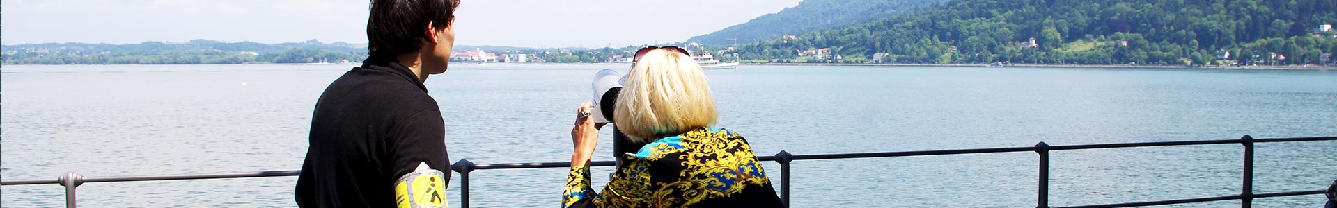 Panoramabild: Eine Frau schaut durch ein Fernglas auf den See und ein Mann mit Blindenschleife steht daneben.